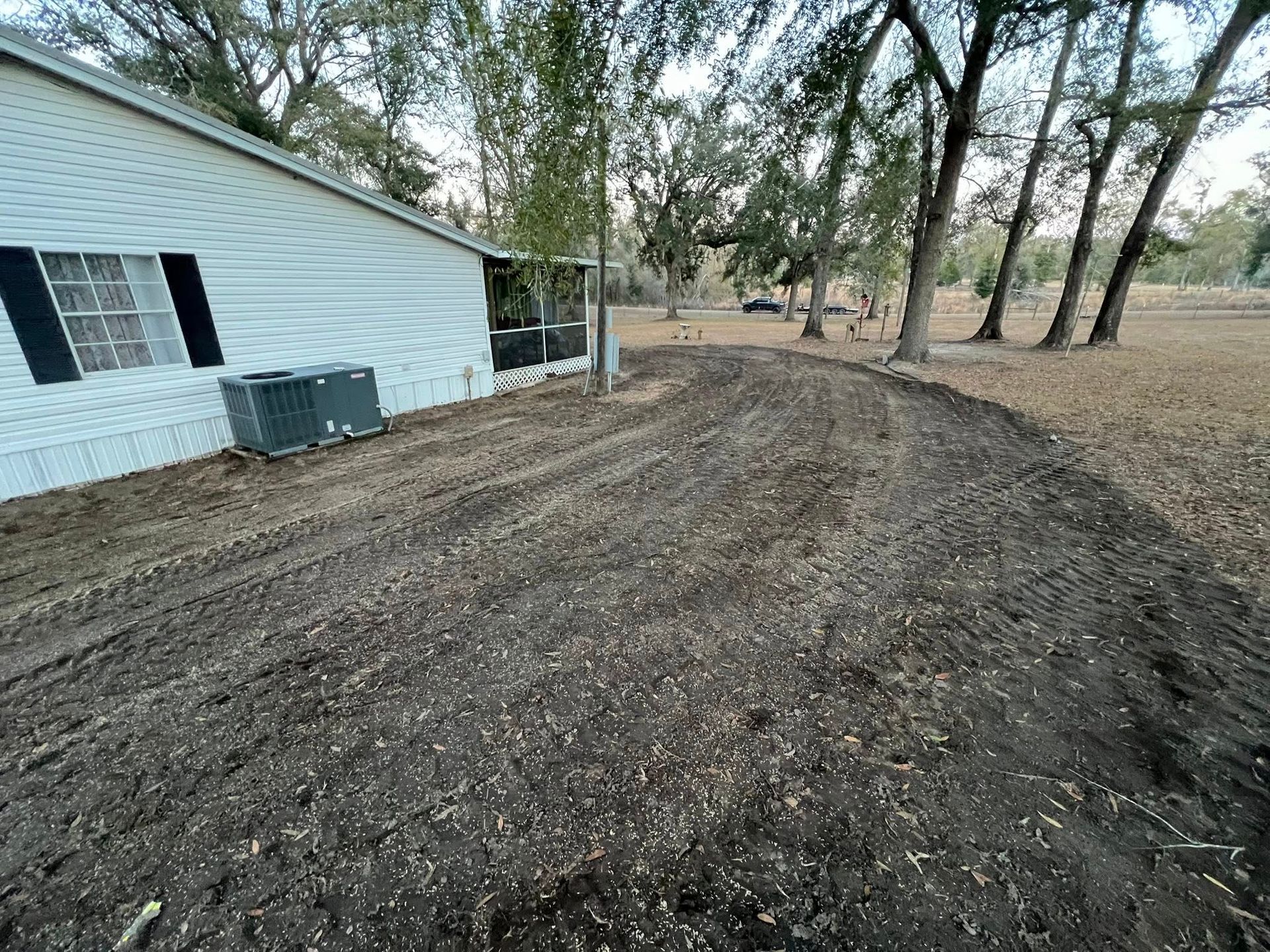 A white mobile home with a dark air conditioning unit next to a newly cleared, bare dirt yard with trees in the background.