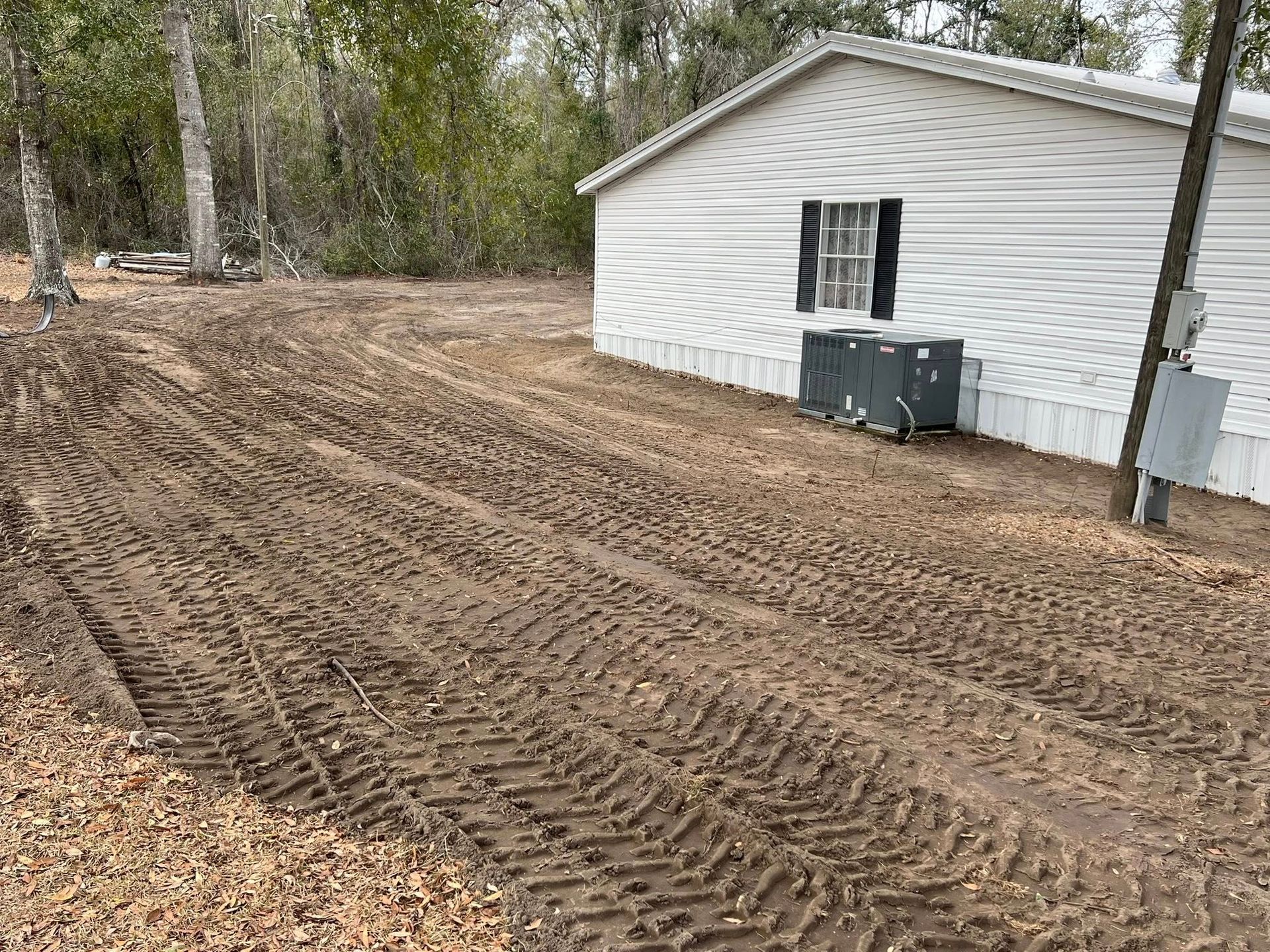 A side view of a white mobile home with a HVAC unit outside, adjacent to a dirt yard with deep, fresh tractor tire tracks.