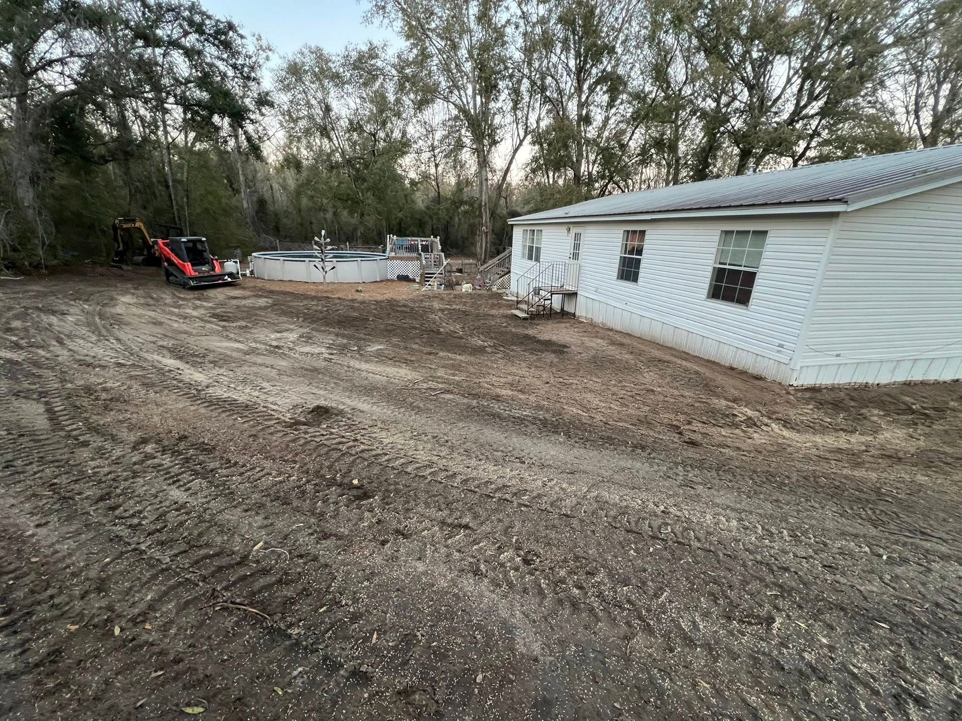 A white house sits next to a dirt yard with tire tracks, a pool frame, and a small red tractor in the background.
