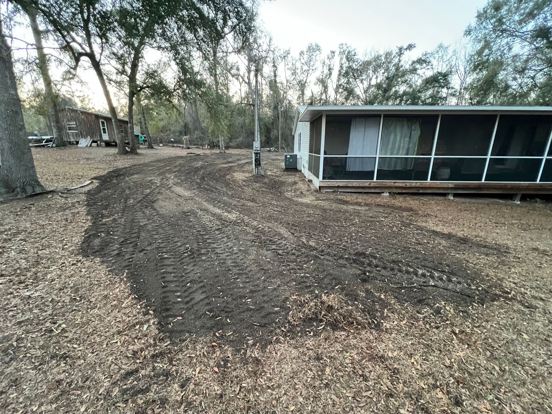 Freshly graded dark dirt patches with tire tracks in a wooded yard beside a house with a screened-in porch.