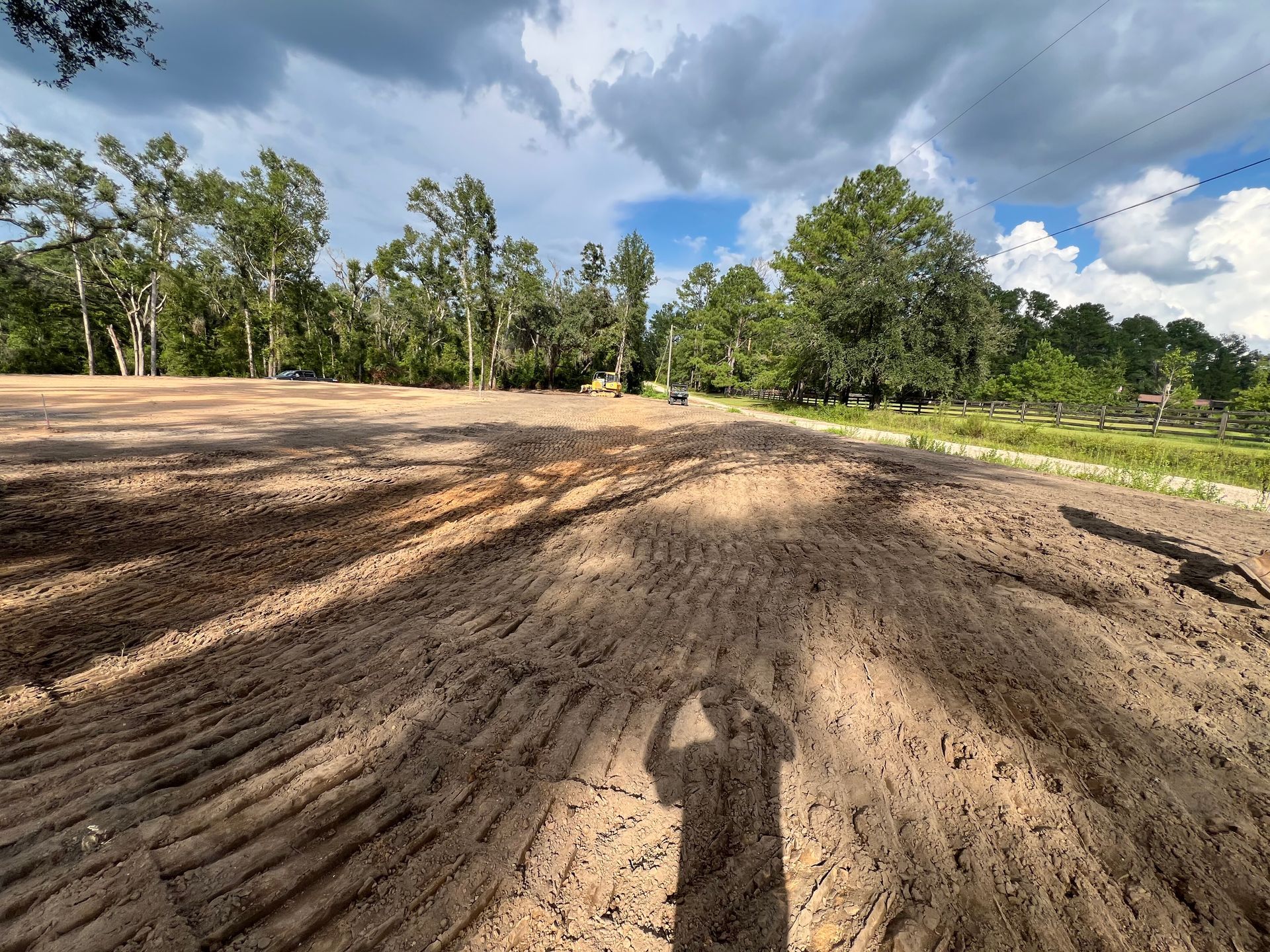 A wide, freshly graded brown dirt field with deep tire tracks, framed by green trees under a cloudy blue sky.