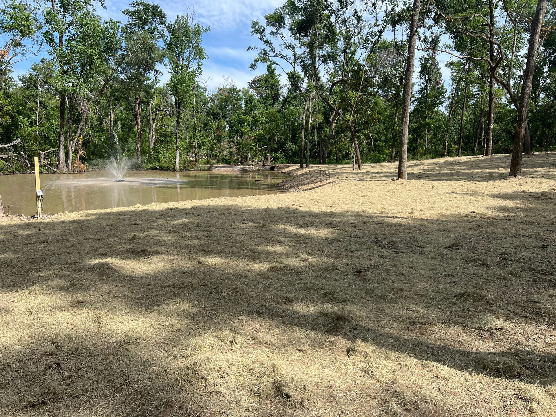 A pond with a fountain sits beside a field of freshly mowed, light brown grass, surrounded by green trees under a blue sky.