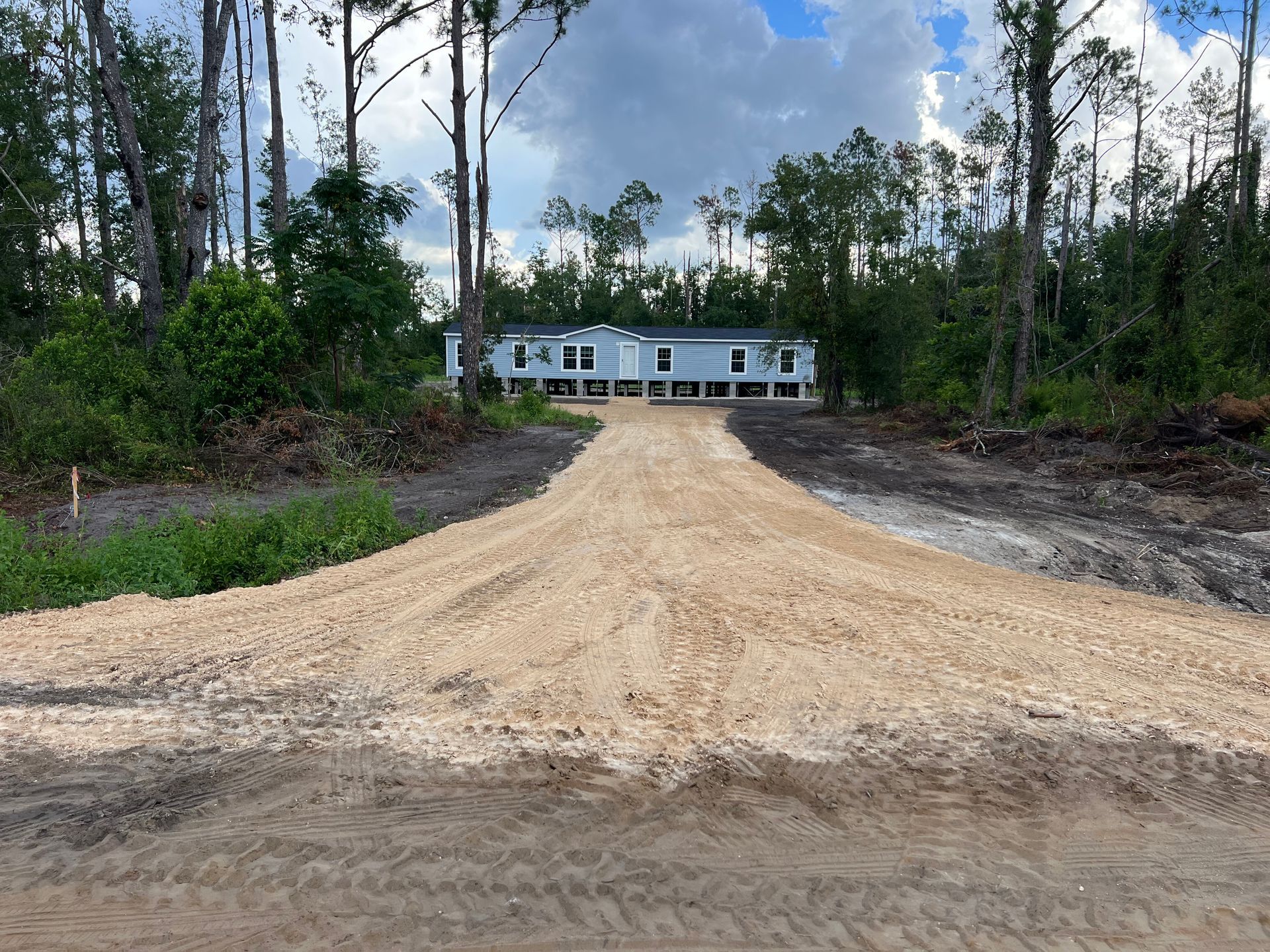 A light blue manufactured home sits at the end of a long, light-colored gravel driveway, surrounded by a wooded lot.