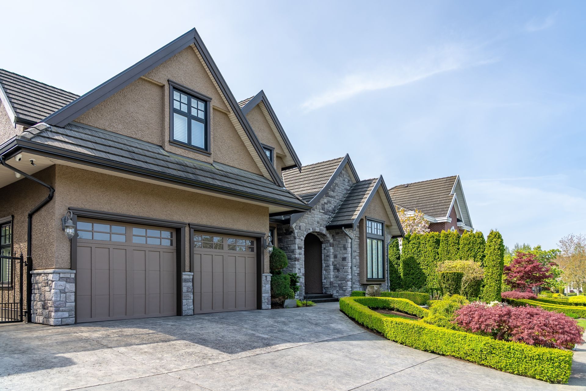 A multi-story suburban house with a brown garage, stone accents, a landscaped garden, and a clear blue sky.