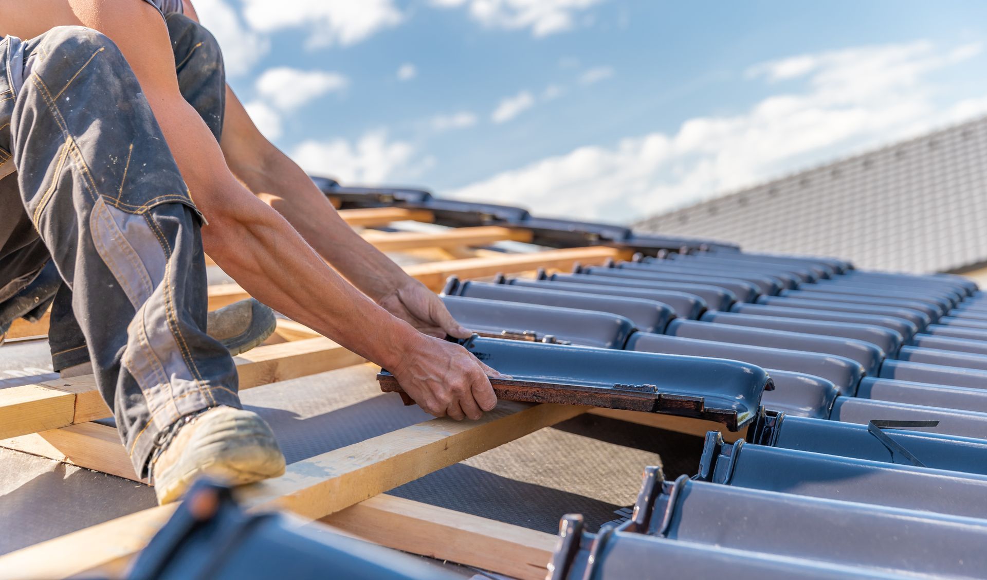 Un ouvrier du bâtiment installe des tuiles sombres et emboîtables sur des liteaux de toit en bois par une journée ensoleillée.