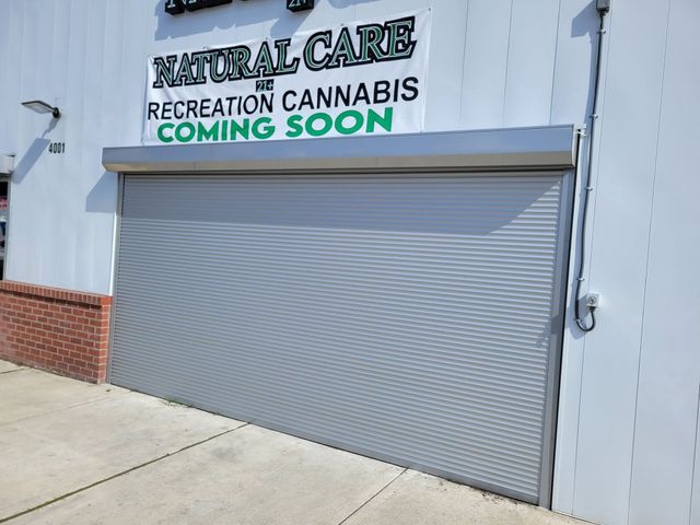 Closed storefront with a gray roll-up door and sign reading “Natural Care Recreation Cannabis Coming Soon.”