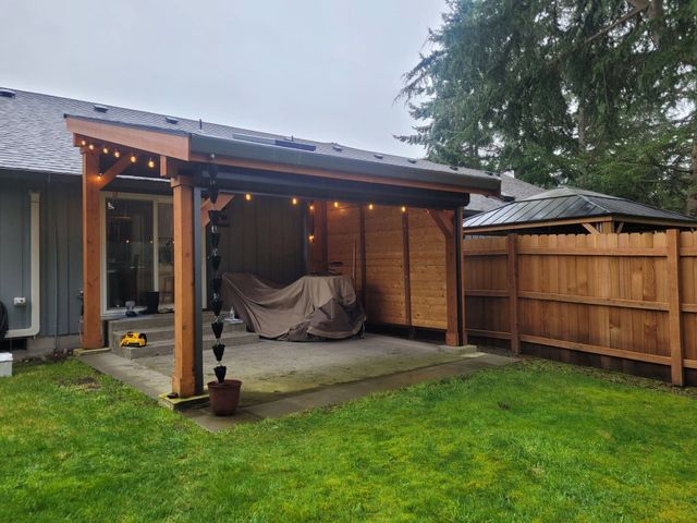 Covered backyard patio with string lights, couch under a tarp, and a wooden fence beside green lawn.