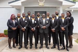 Eight people in formal black suits stand posing outside a house with a white porch.