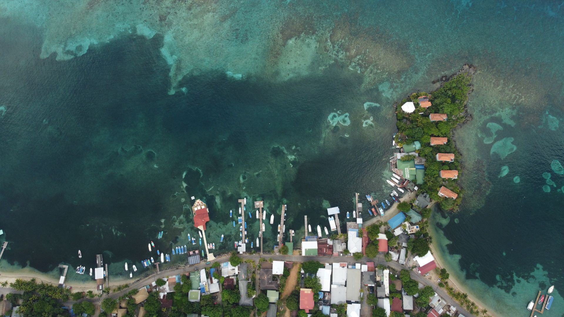 Aerial view of a coastal village with boats docked at a pier, turquoise water surrounds the land, and a small island with cabins.