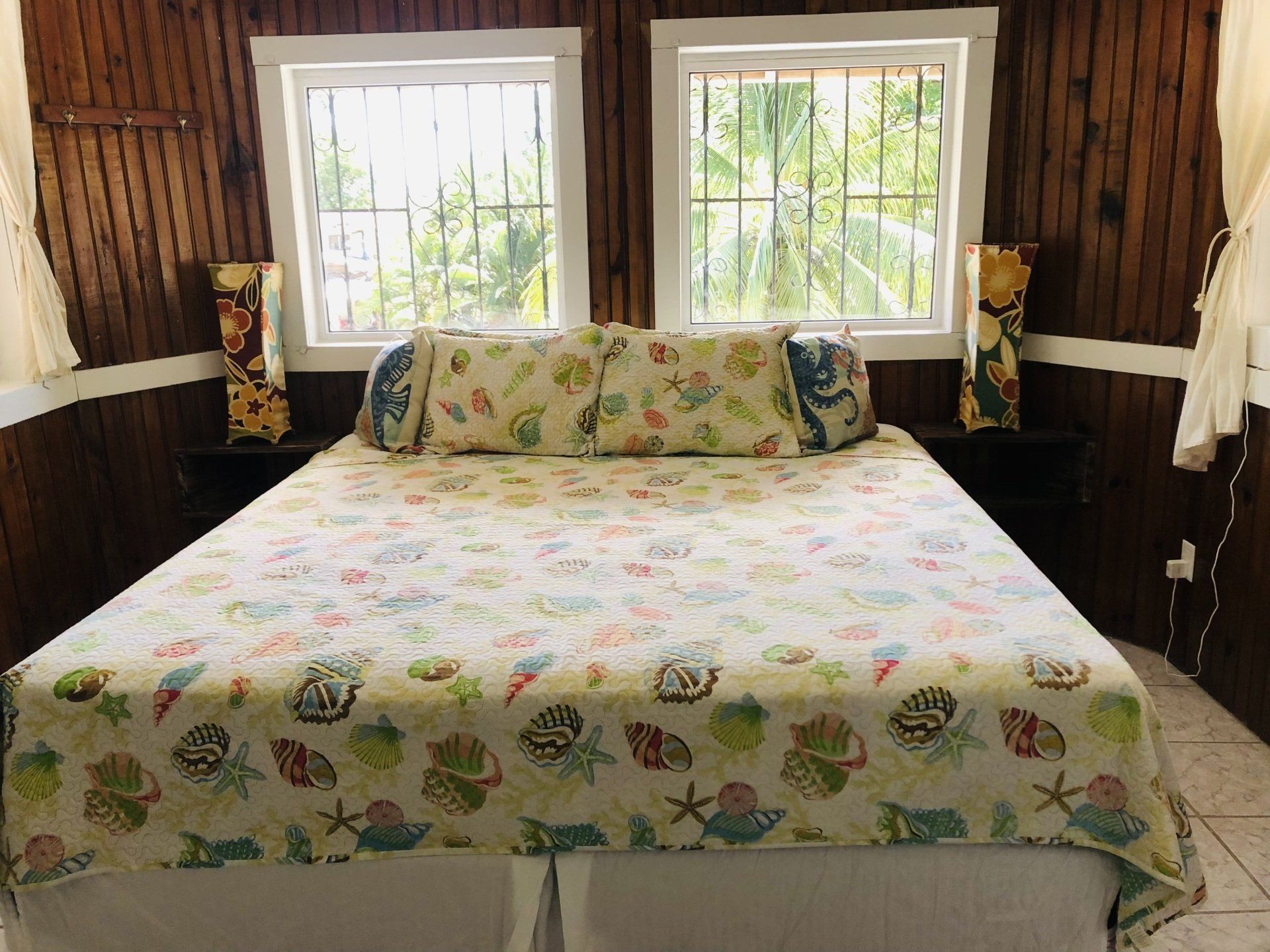 Bedroom with seashell-patterned bedding, wooden walls, two windows, and decorative lamps on black nightstands.