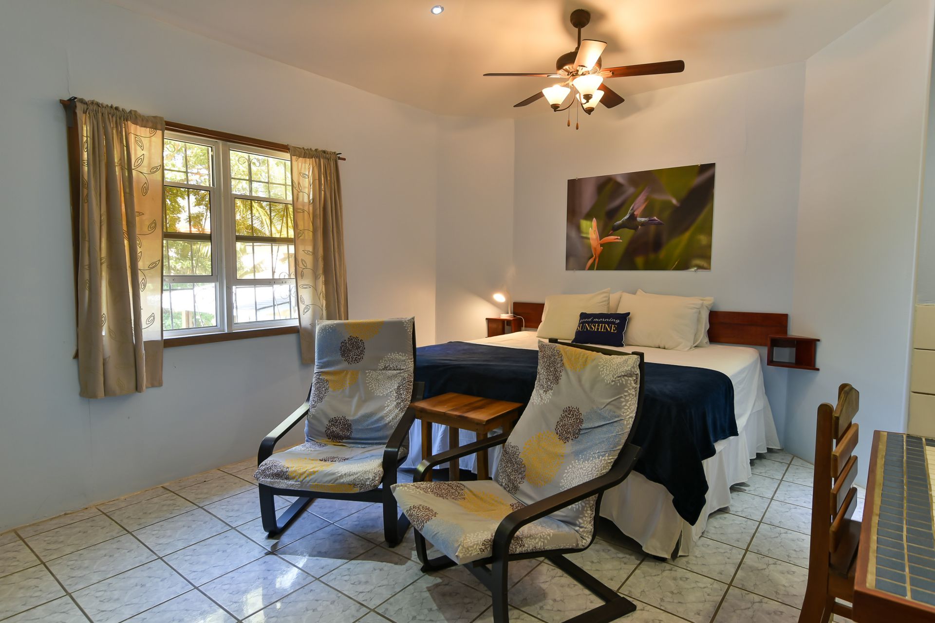 Bedroom with a queen bed, two chairs, a window with curtains, and artwork; bright room.