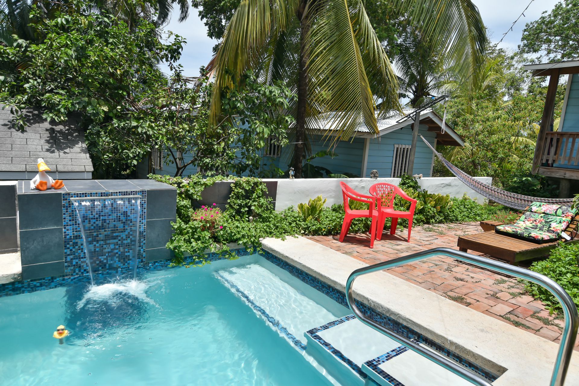 Pool with waterfall, red chairs, hammock, and lush greenery in outdoor setting.