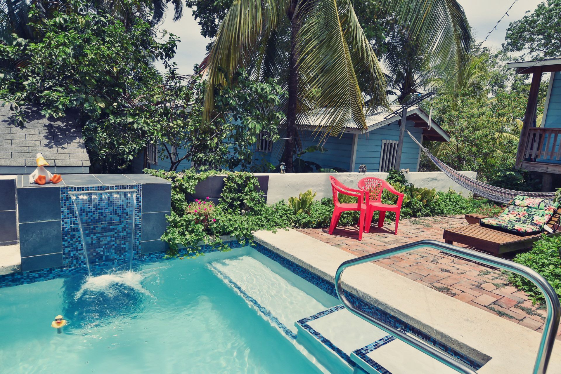 Poolside scene: turquoise water, small waterfall, red chair, hammock, and lush greenery.