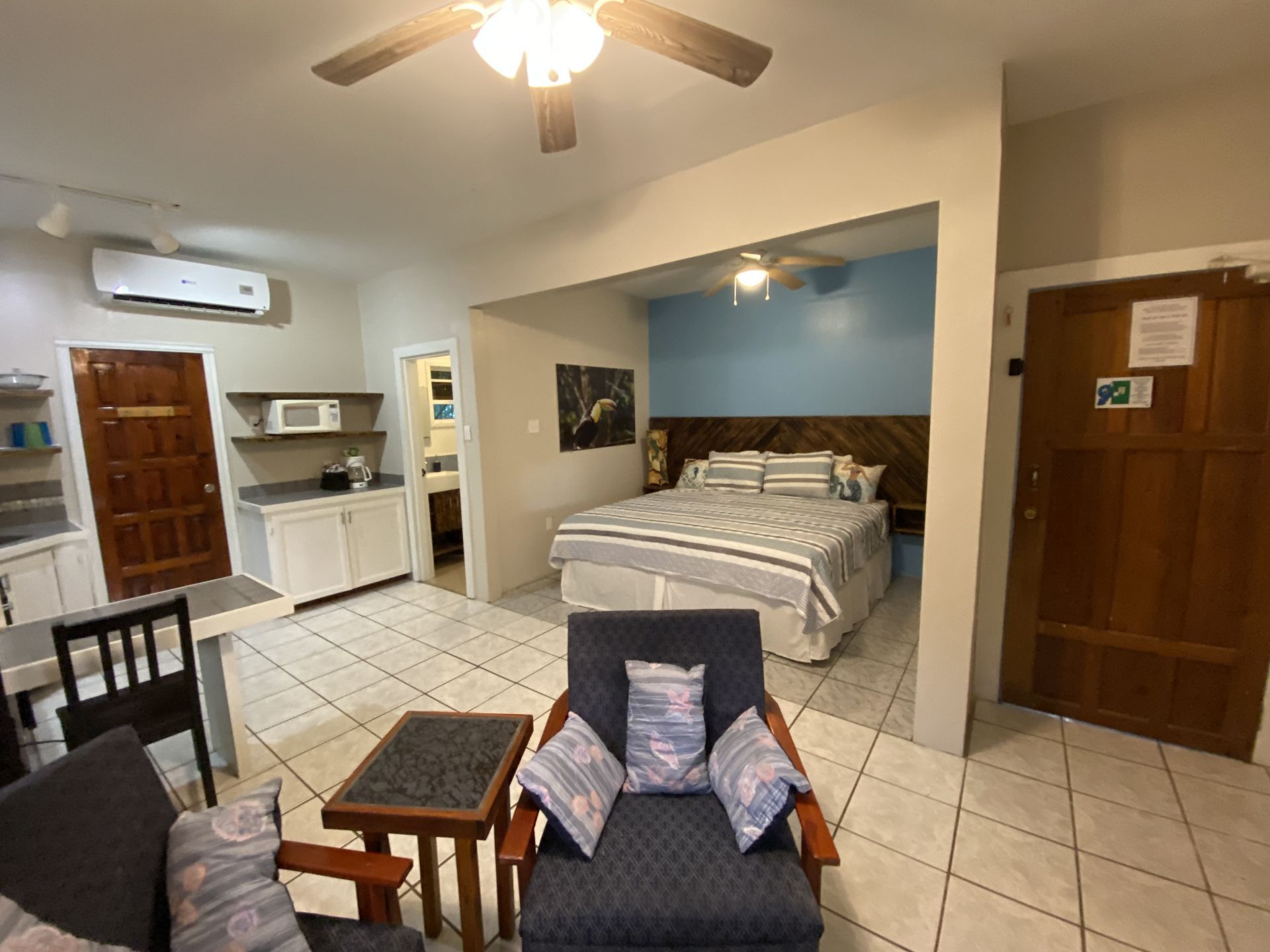 Studio apartment interior: bed, seating, kitchen area, and bathroom access. White tile floor, blue accent wall.