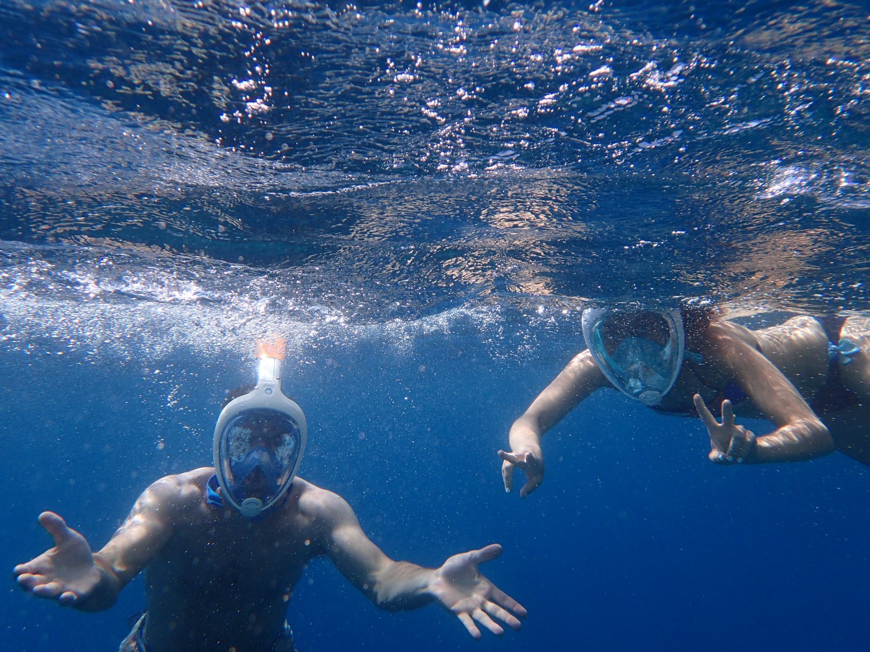 Two snorkelers underwater in blue water, arms outstretched, wearing full-face masks, one making a peace sign.