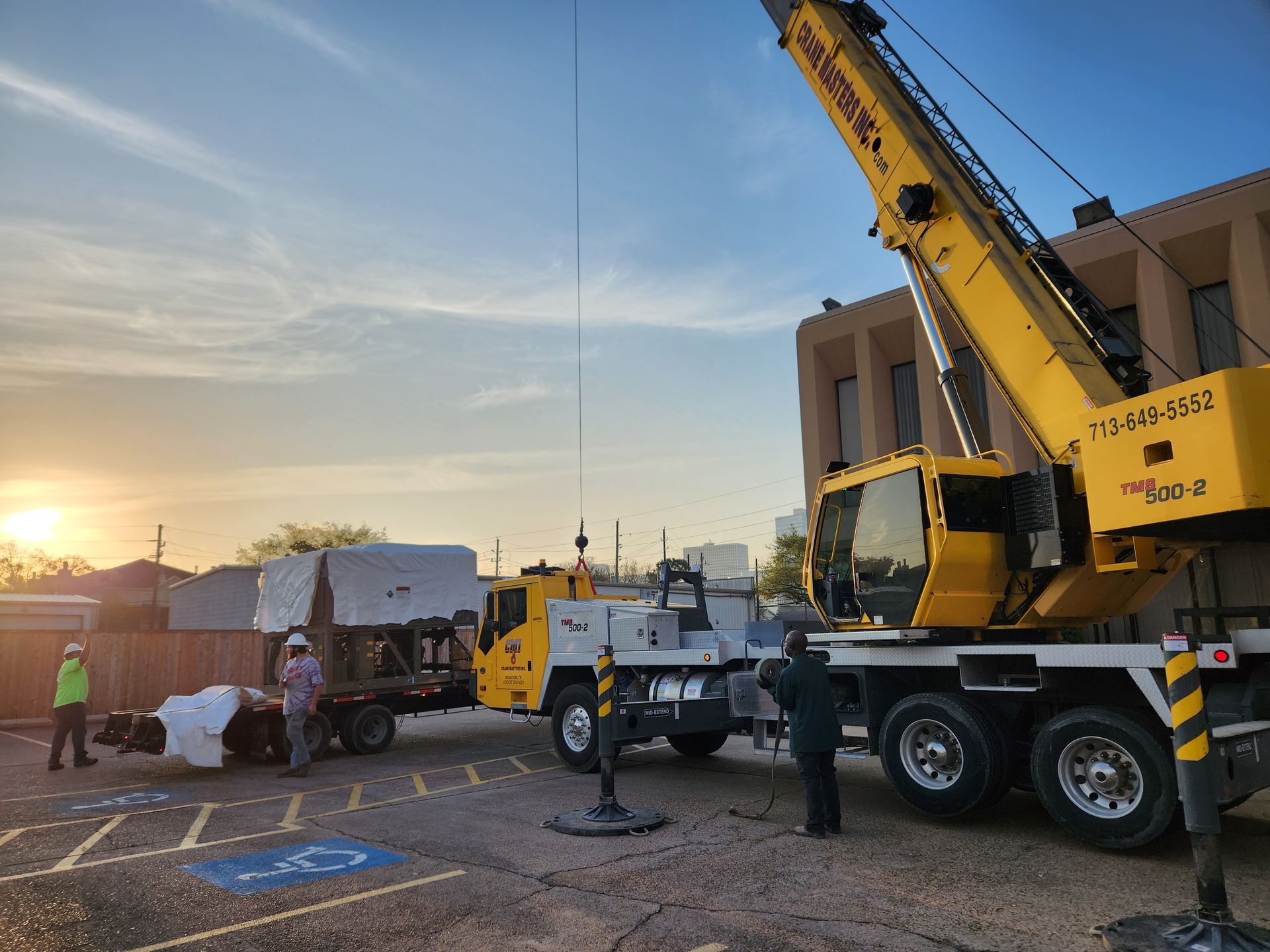 Crane lifting equipment on a truck at sunset in a parking lot, with workers nearby