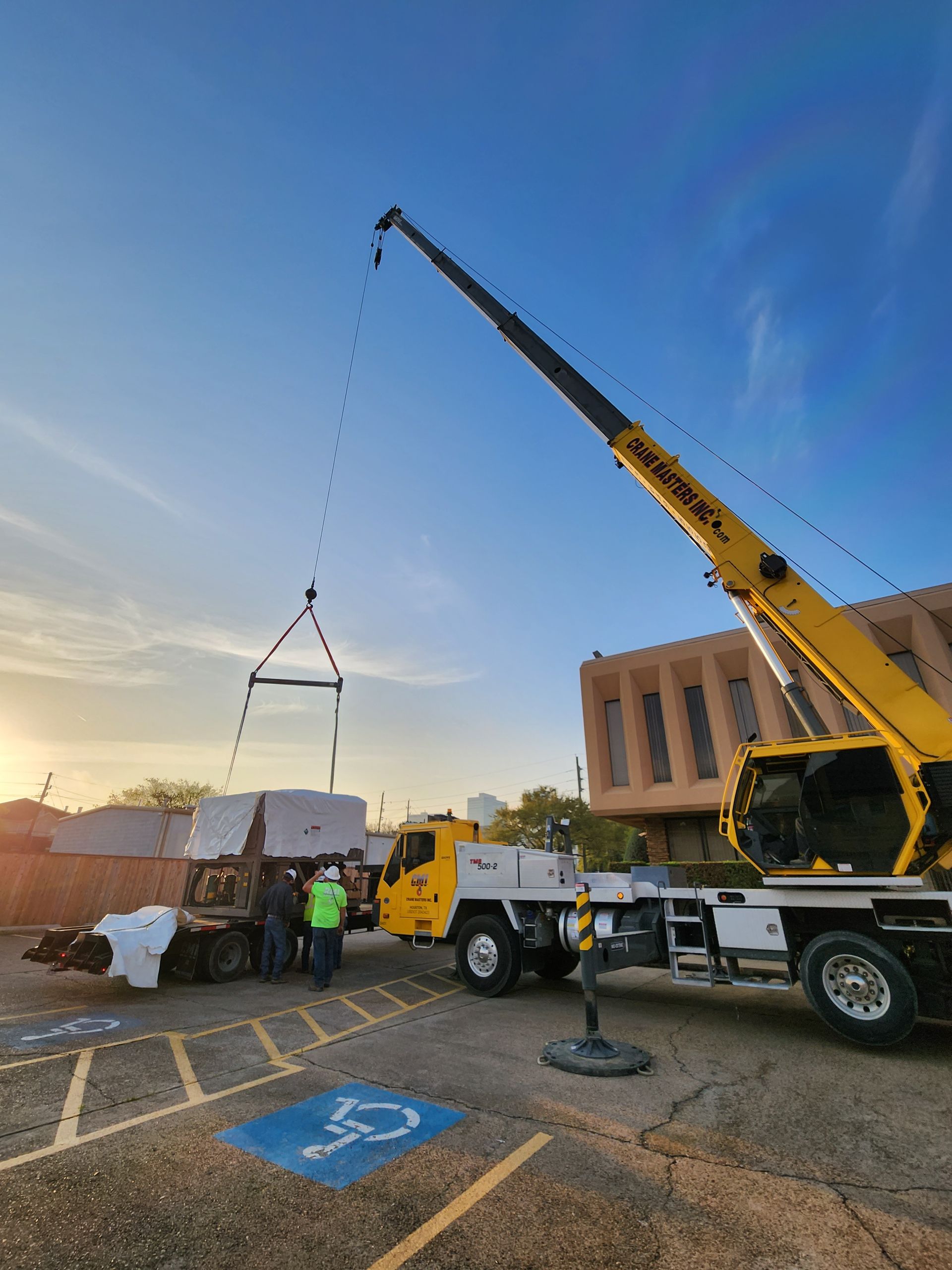 Crane lifting a white structure at dusk in a parking lot beside a yellow truck and building