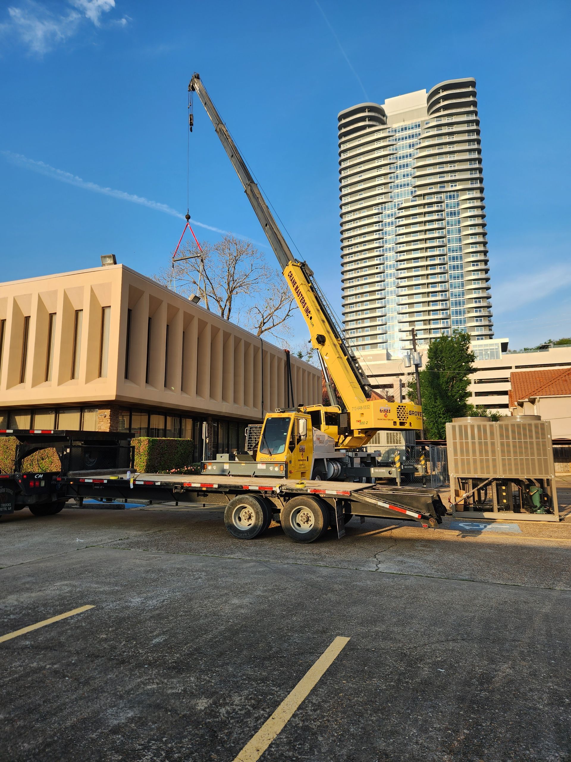 Construction crane lifting a large beige building module at a worksite, with a tall tower in the background.