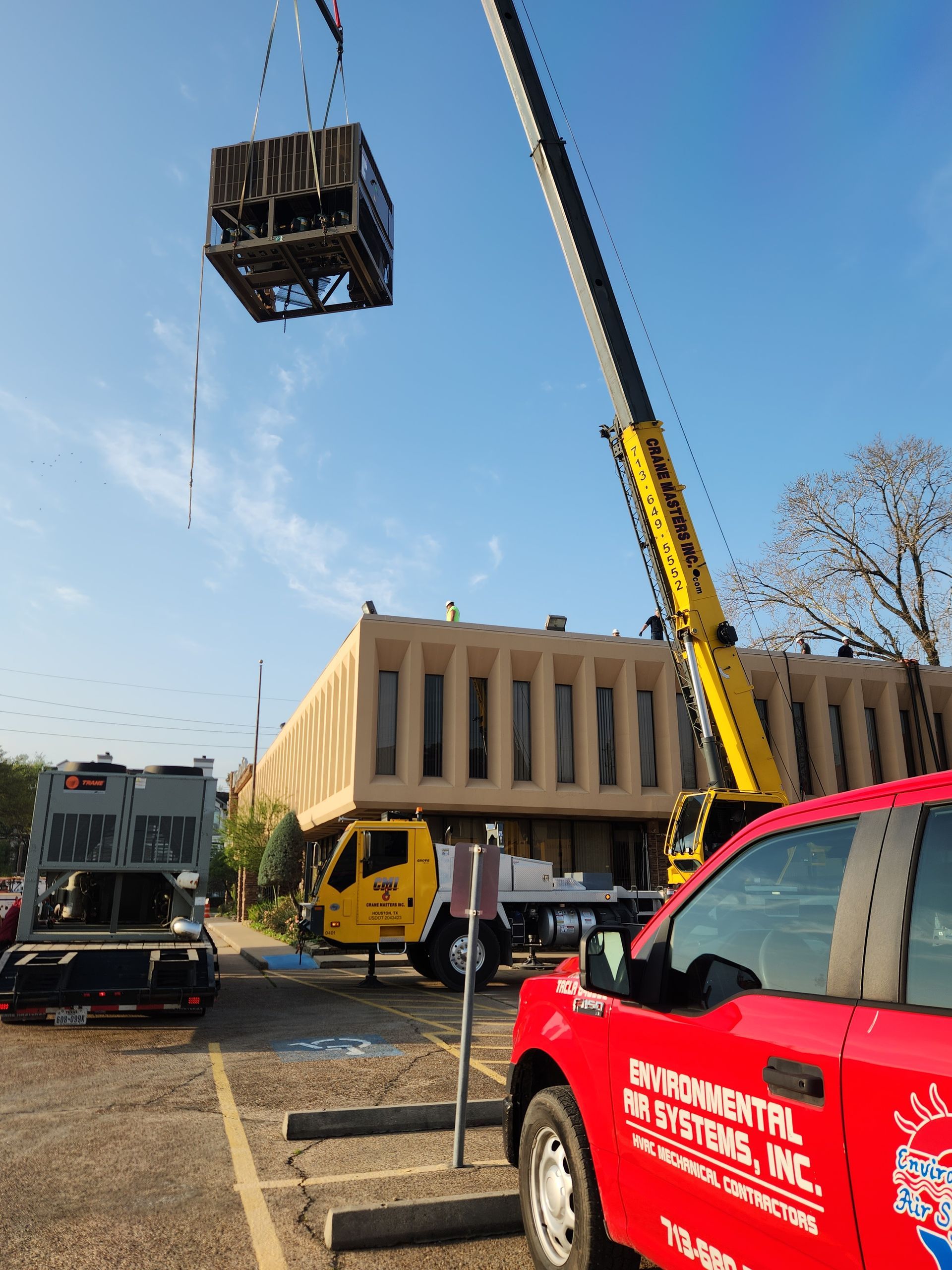 Crane lifting a large black crate beside a beige building, with red and yellow vehicles in the foreground.
