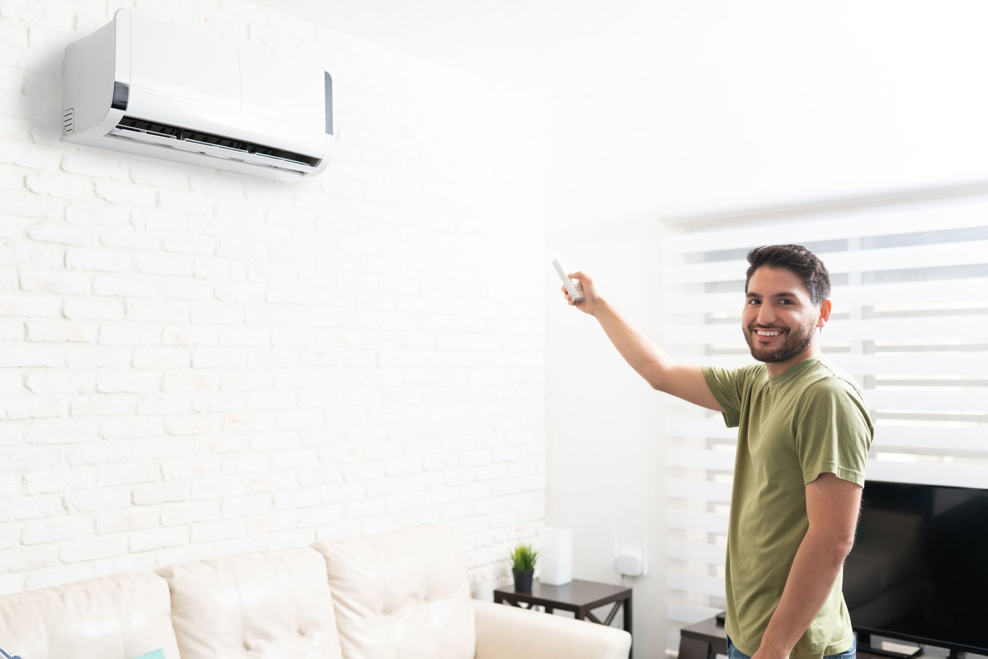 Smiling man using a remote to control a wall-mounted mini-split AC unit in a modern living room. Smiling man using a remote to control a wall-mounted mini-split AC unit in a modern living room.