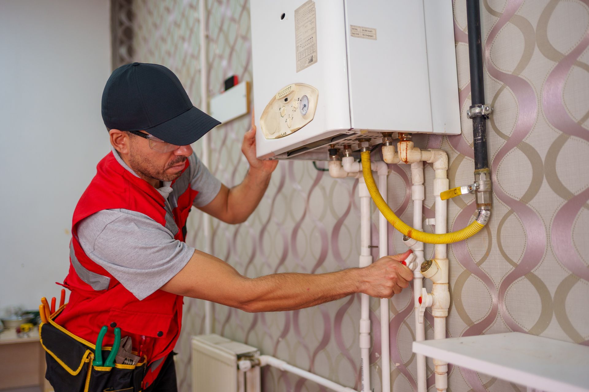 A technician wearing a red vest and hat carefully works on a residential heater installation.