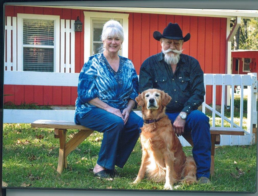 Two people sit on a bench with a golden retriever in front of a red house.