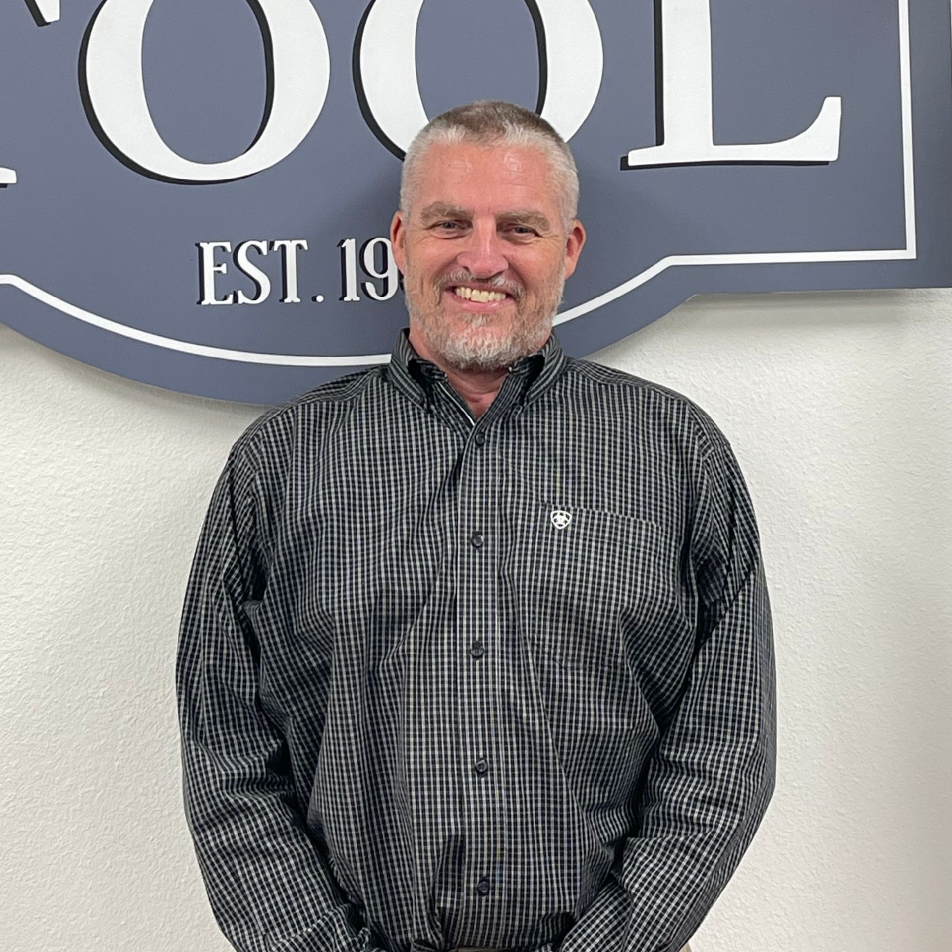 Smiling man in a dark plaid shirt standing in front of a wall sign