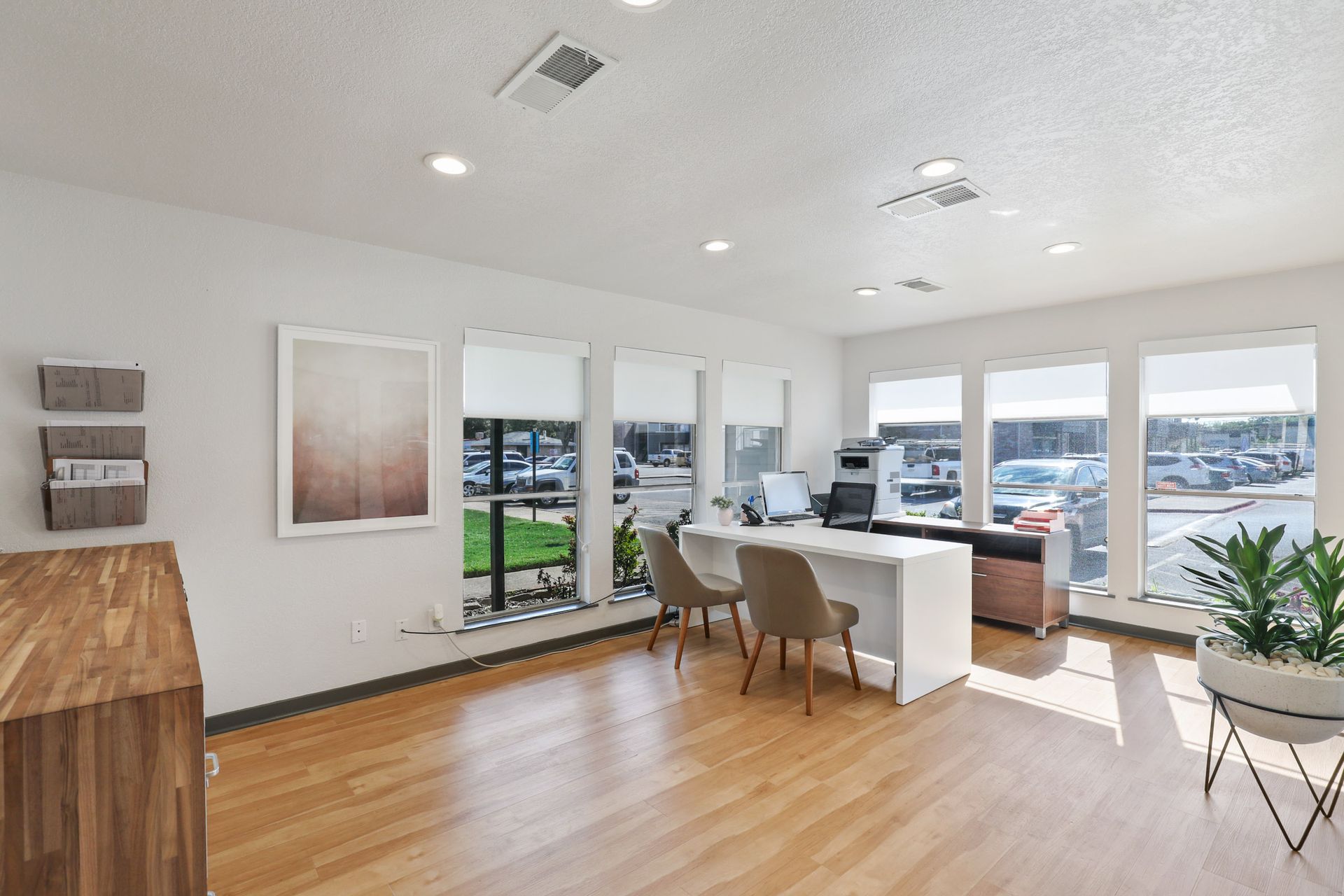 Bright office with desk, chairs, large windows overlooking outdoor area, hardwood floors, and potted plants.