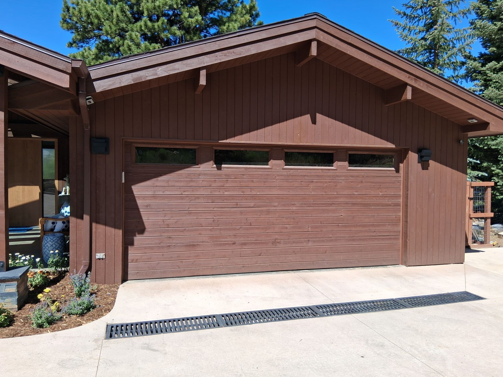 A wooden garage with a four-paneled door, brown siding, and a long horizontal drainage grate on the concrete driveway.