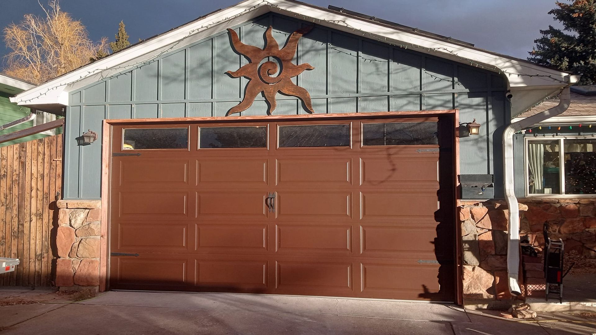 A brown garage door with windows, decorated with a sun wall art piece on blue siding, flanked by stone walls.