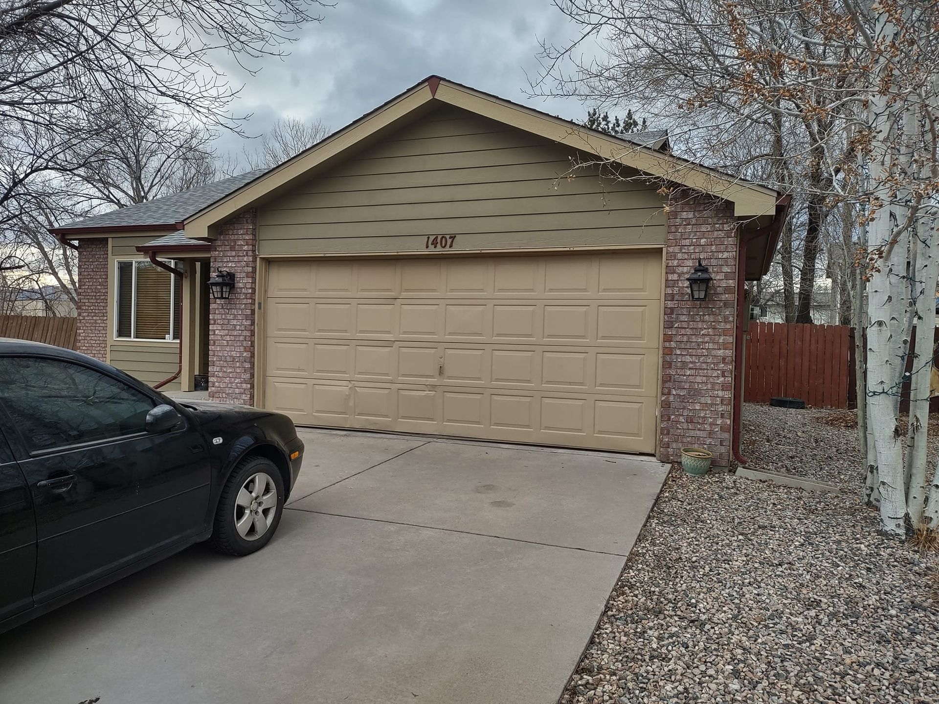 A beige, single-story residential garage with brick accents, seen from the driveway with a black car parked to the left.