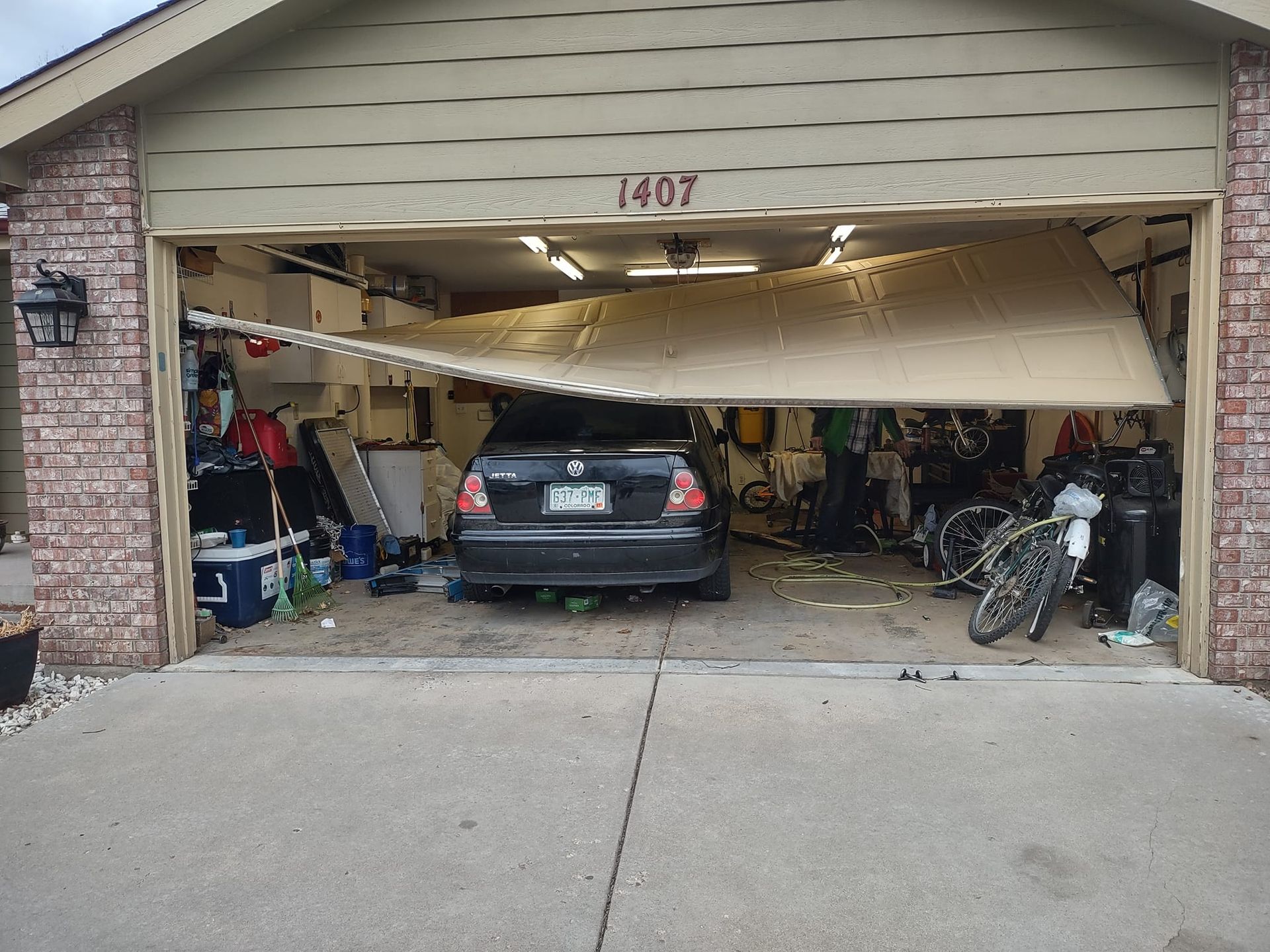 A residential garage door has come off its tracks and is hanging at an angle in front of a parked black sedan.