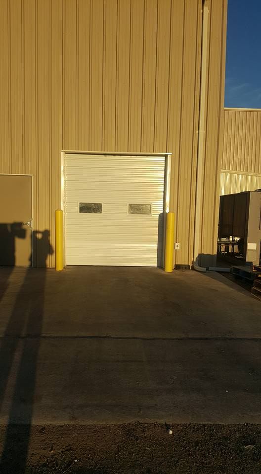 A metal warehouse building with a closed rolling garage door, flanked by two yellow safety bollards on a concrete lot.