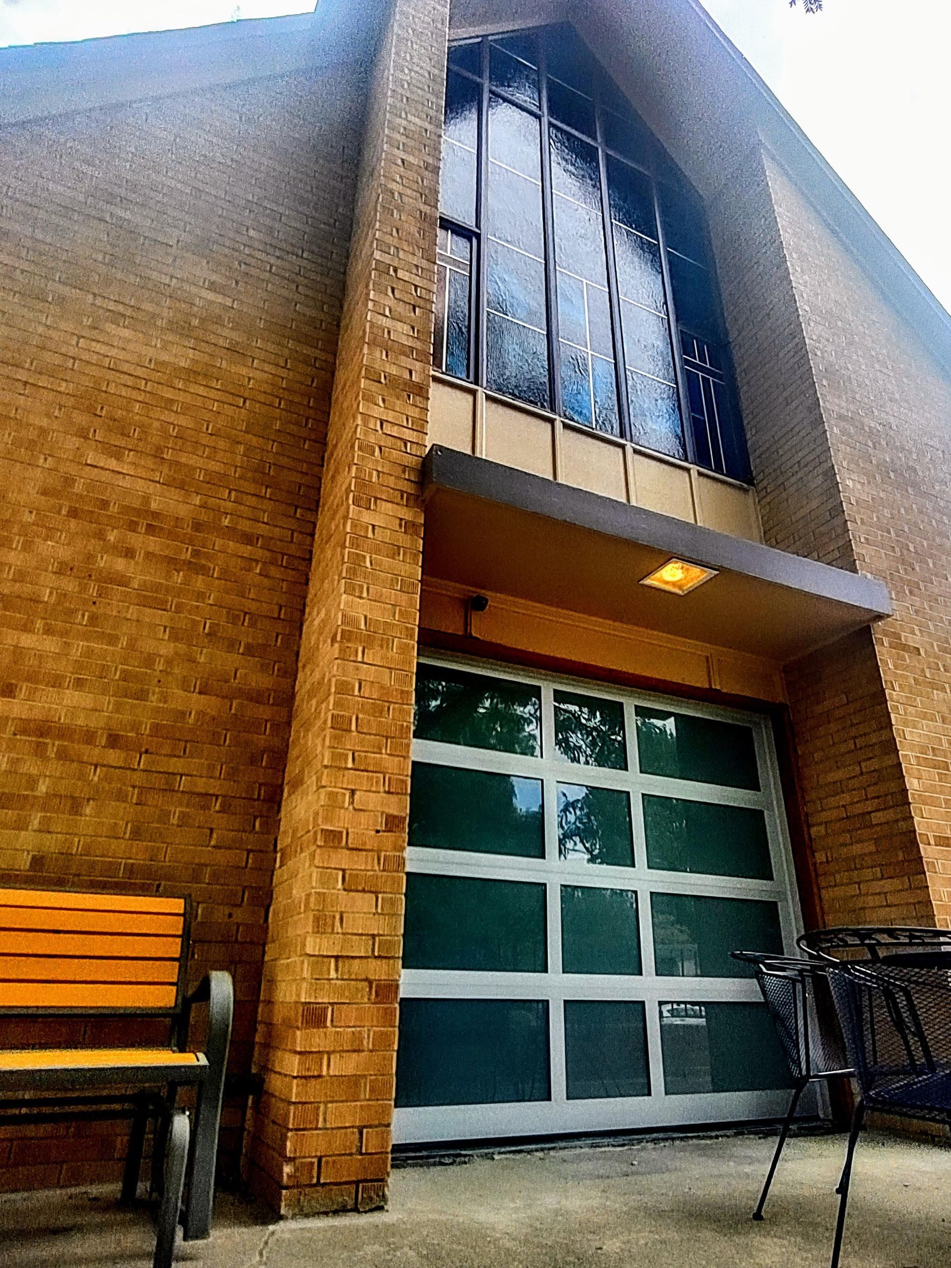 A low-angle view of a brick building entrance with a modern, glass-paneled garage-style door below a tall stained-glass window.