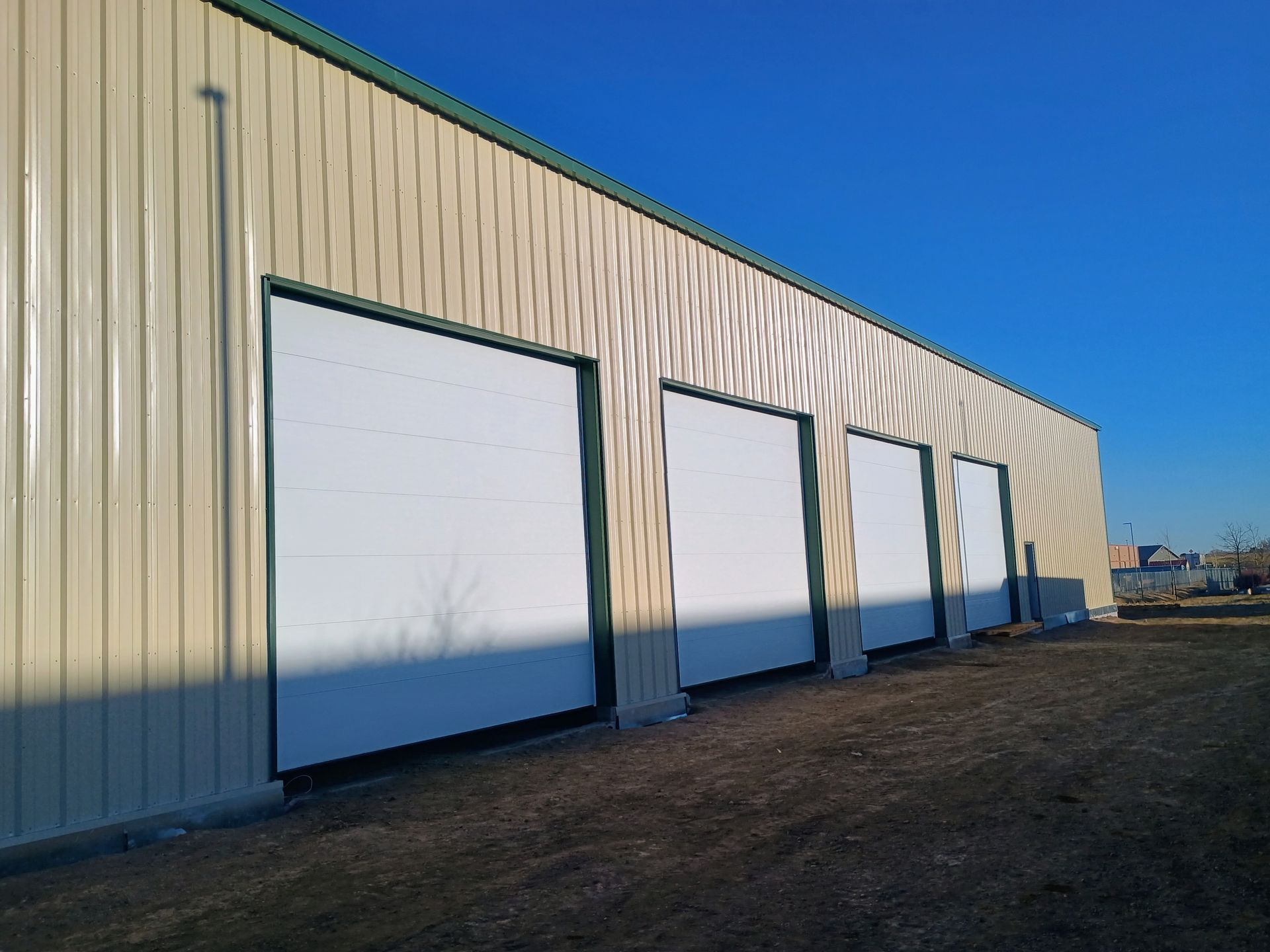A tan metal industrial building with four white roll-up doors set against a clear blue sky.