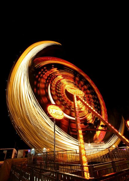 A brightly lit, spinning carnival ride at night, creating circular light streaks against a dark sky.
