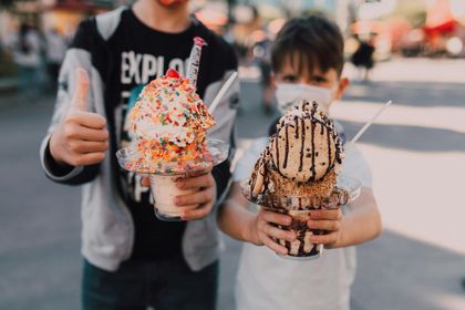 Two people holding large, elaborately decorated ice cream sundaes outdoors, one giving a thumbs-up.