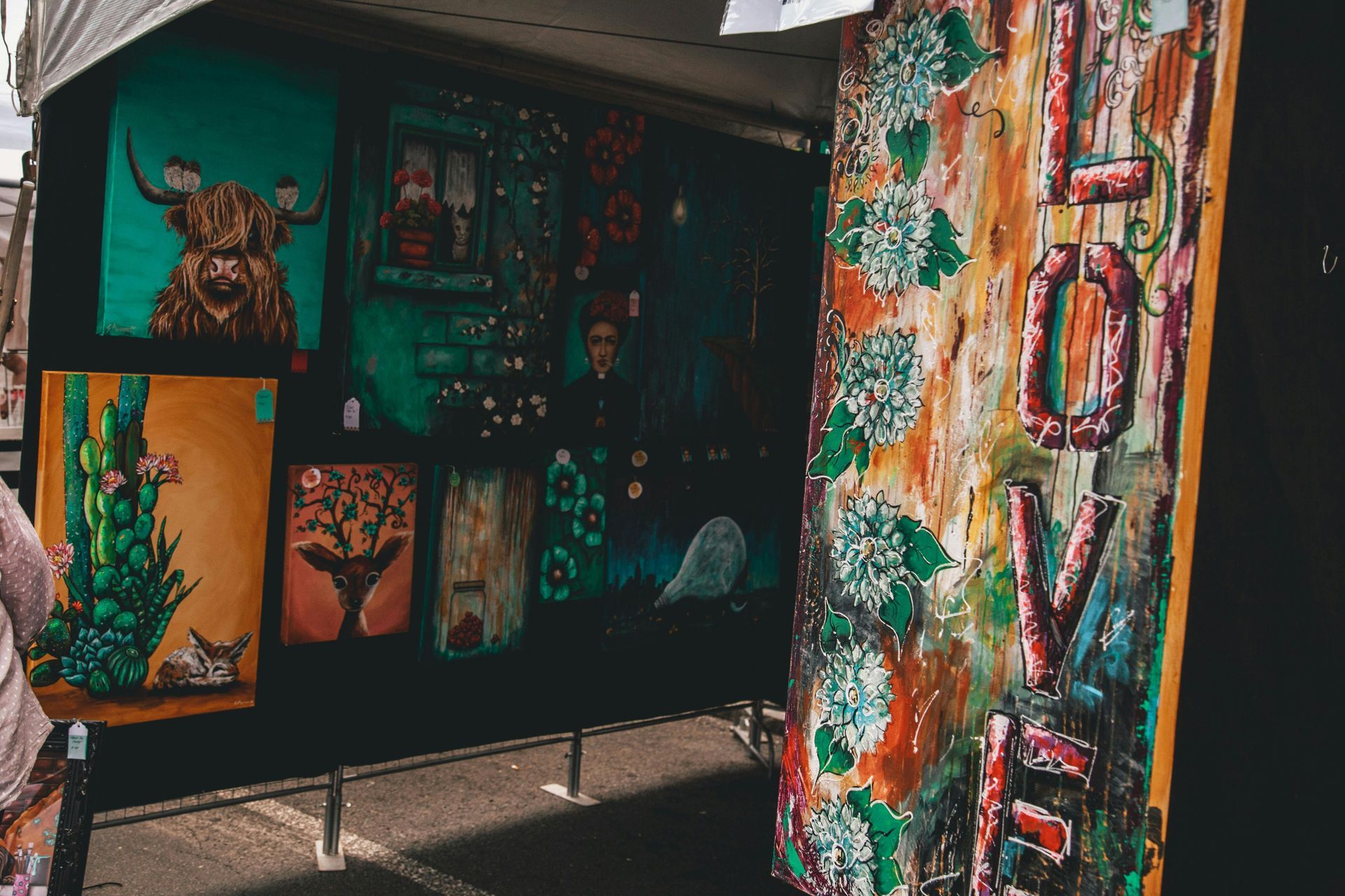 An art display featuring a tall, vertical 'LOVE' sign, various landscape paintings, and a portrait of a highland cow.