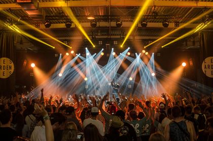 A crowded concert venue with a stage illuminated by bright, dynamic beams of yellow and white light.