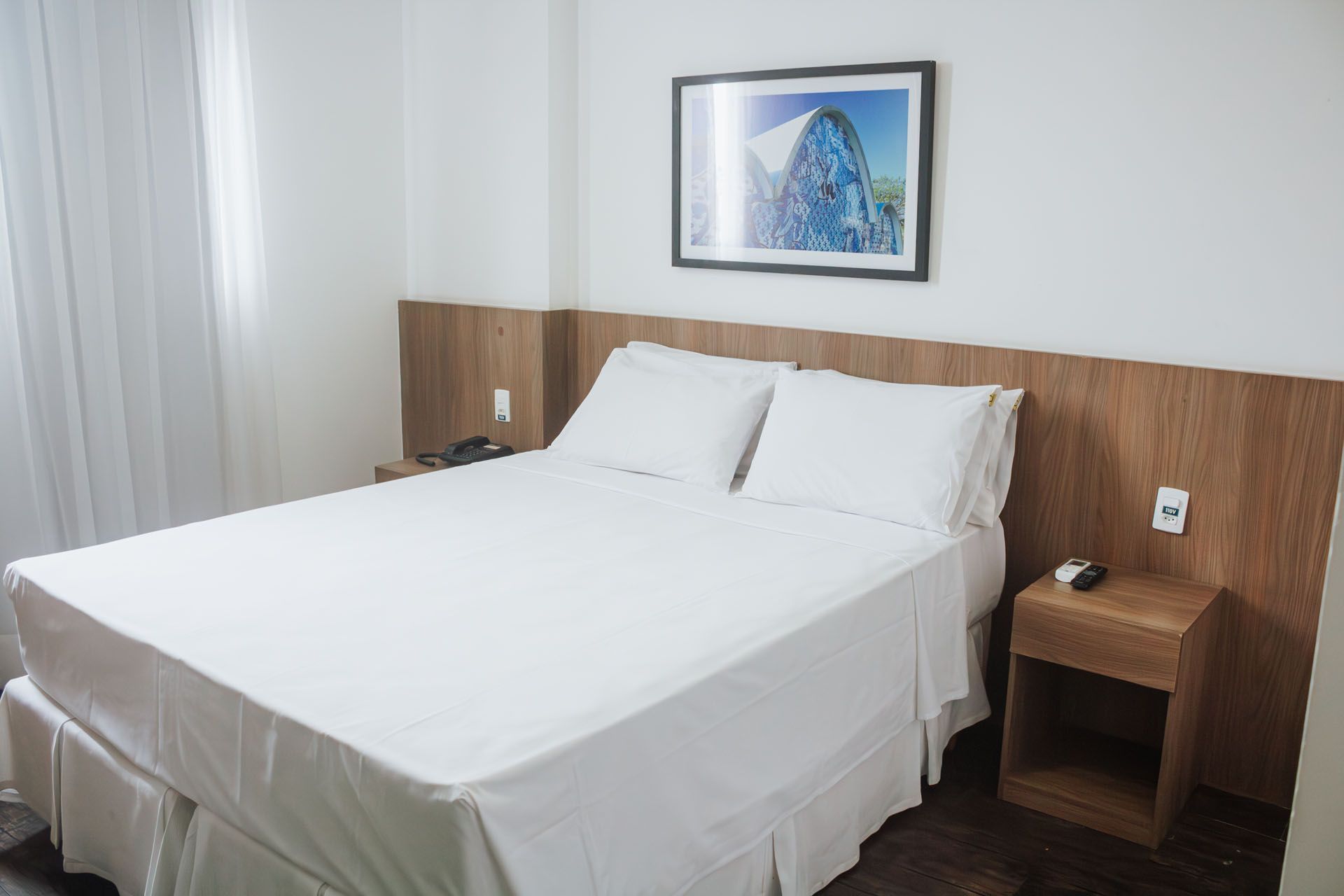 Quarto de hotel com uma cama branca impecavelmente arrumada, cabeceira de madeira, mesa de cabeceira e obras de arte emolduradas na parede.