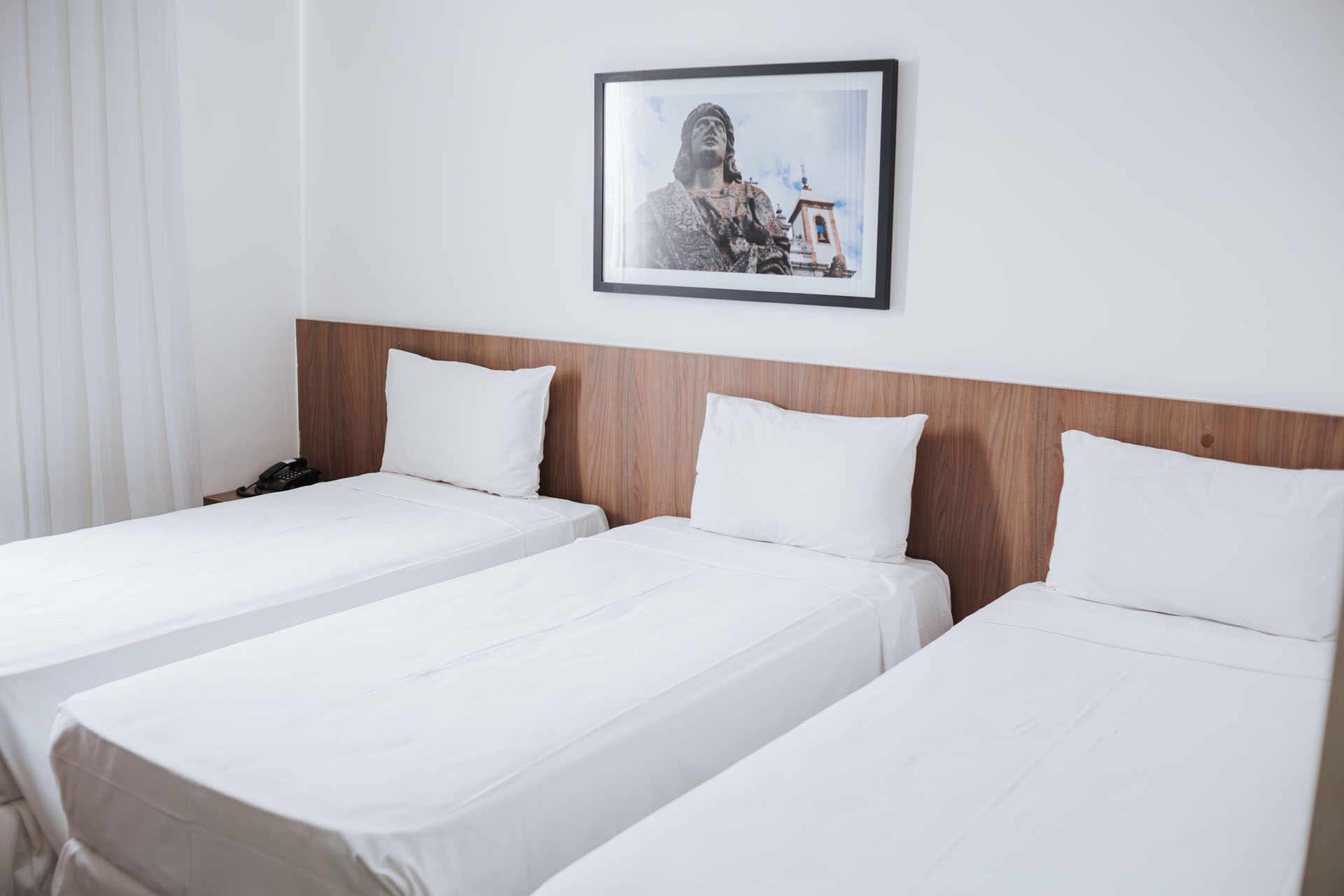 Quarto de hotel luminoso com três camas brancas impecavelmente arrumadas e um quadro acima da cabeceira.