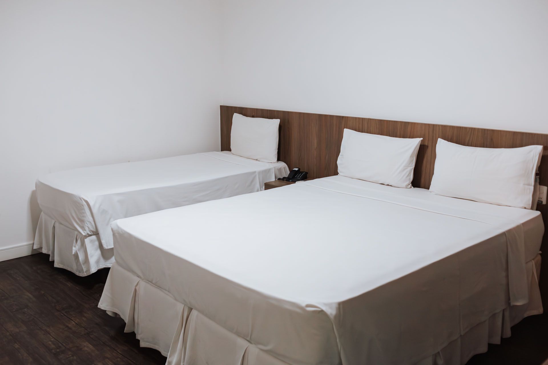 Duas camas brancas impecavelmente arrumadas em um quarto de hotel simples com cabeceiras de madeira.
