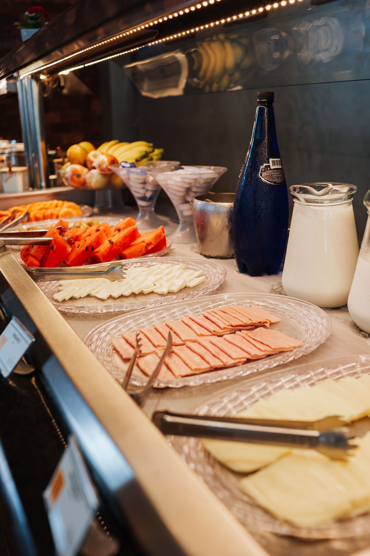 Mesa de buffet com frios fatiados, morangos, iogurtes, garrafas de leite e garrafas de vidro azul sob iluminação aconchegante.