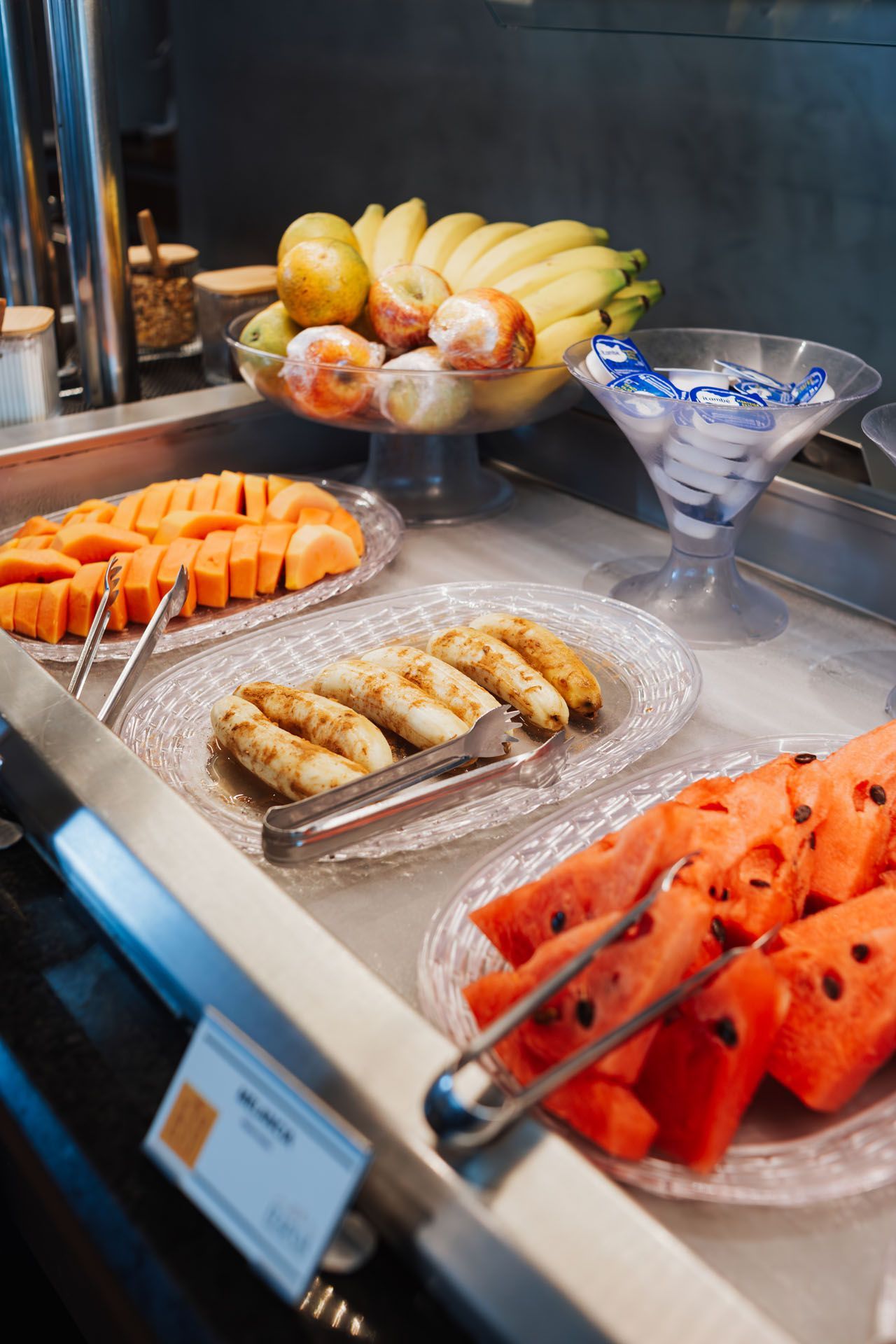 Mesa de buffet com frutas fatiadas, doces e uma tigela de sobremesa azul e branca.