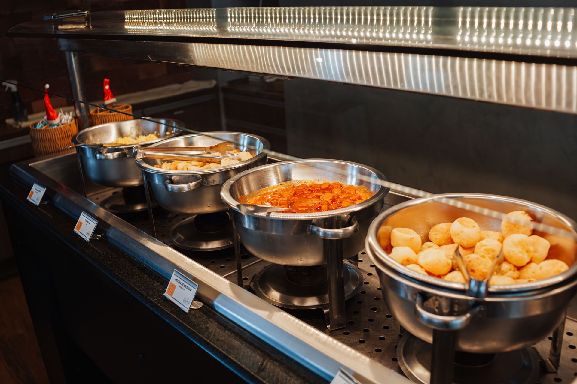 Balcão de buffet com recipientes metálicos contendo comida quente, incluindo massas e pãezinhos, sob uma luz aquecedora.