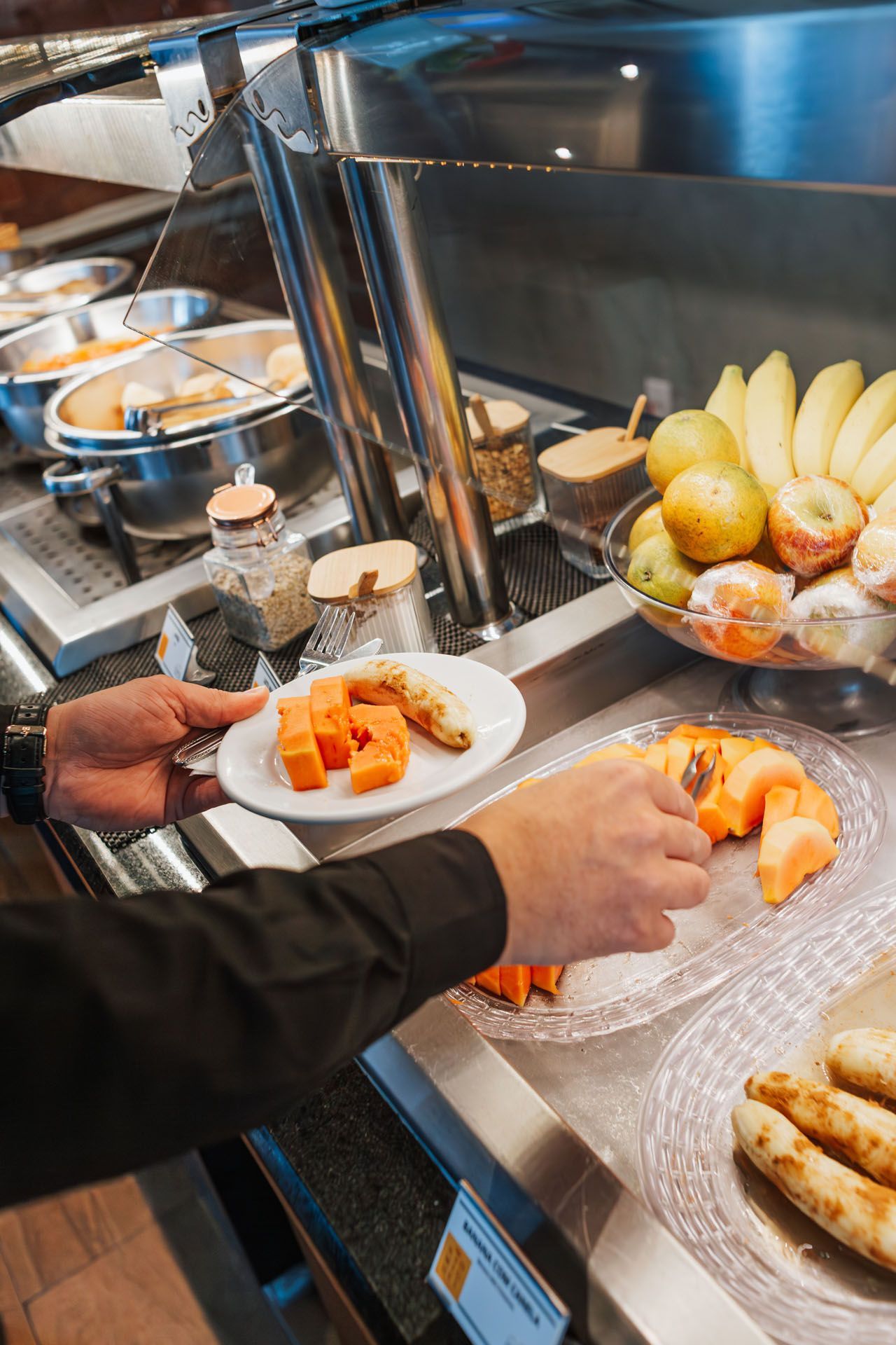 Mãos servindo camarão e frutas em um balcão de buffet em uma área de refeições de aço inoxidável.