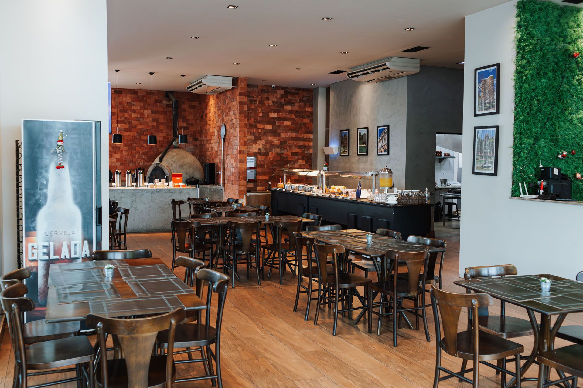 Sala de jantar vazia de um restaurante com mesas de madeira, cadeiras pretas, parede de tijolos ao fundo e balcão.