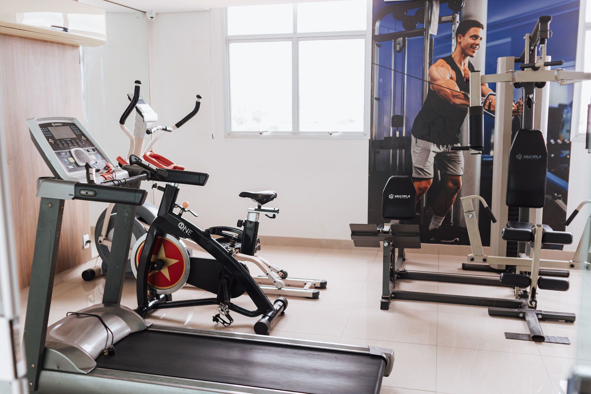 Academia em casa com esteira, bicicleta ergométrica, pesos e um homem usando um aparelho de musculação.