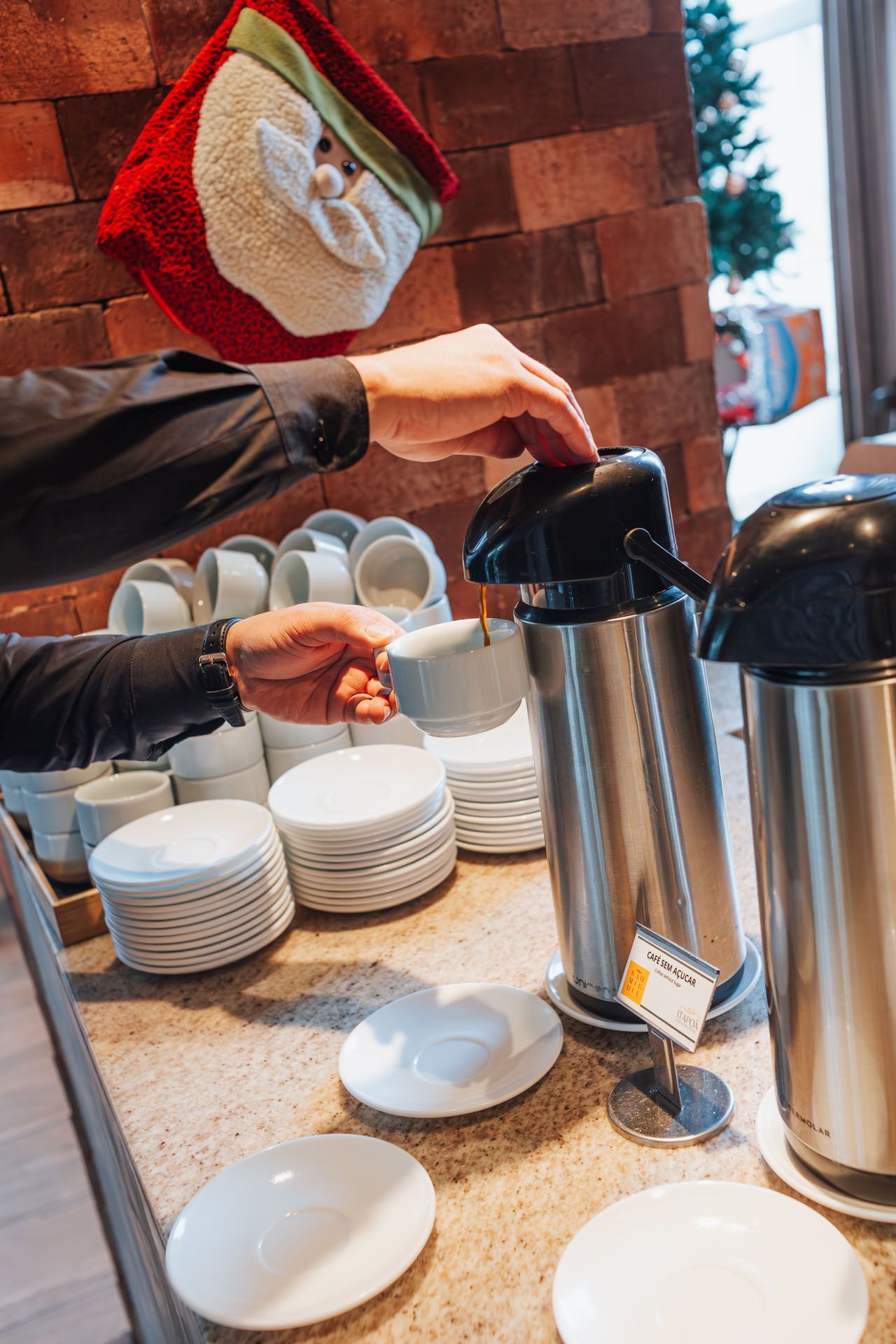 Mãos servindo café de uma garrafa térmica para uma xícara branca em um buffet com pratos empilhados e garrafas térmicas.
