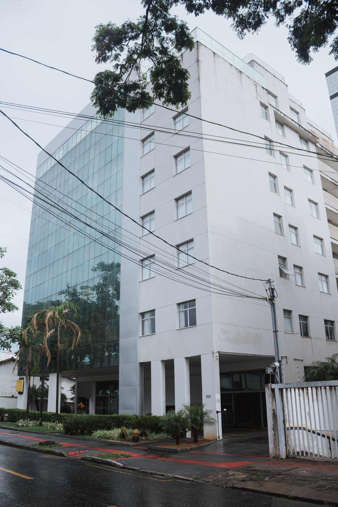 Prédio alto de escritórios, branco e de vidro, em uma rua chuvosa, visto de baixo com árvores acima.